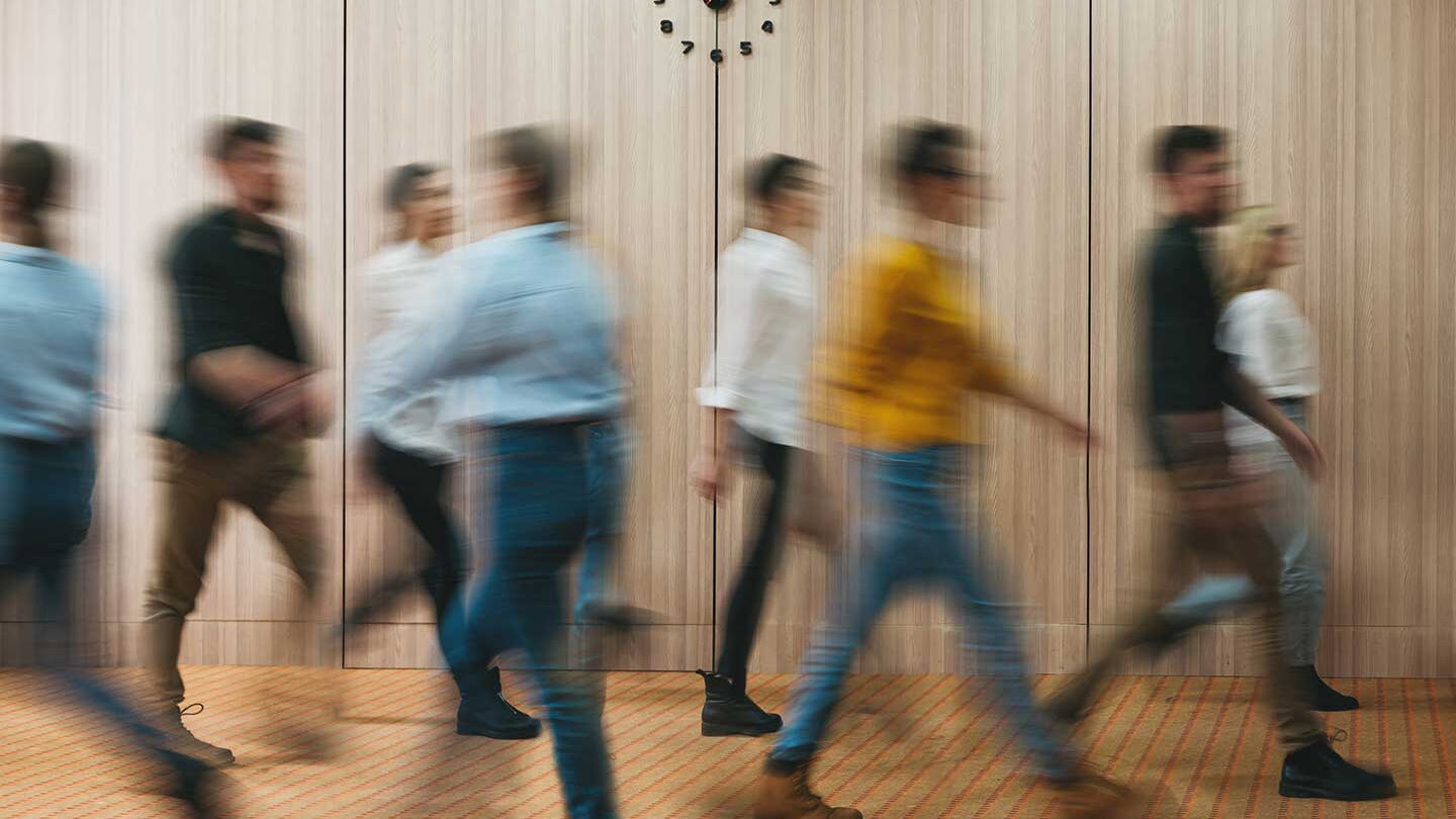 Acht Männer und Frauen gehen kreuz und quer durch einen braun getäfelten Gang.