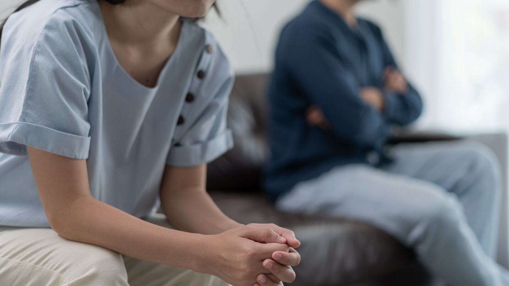 Im Vordergrund sitzt eine Frau auf einem Sofa und stützt die Ellbogen auf die Oberschenkel ab. Im Hintergrund sitzt ein Mann mit verschränkten Armen.