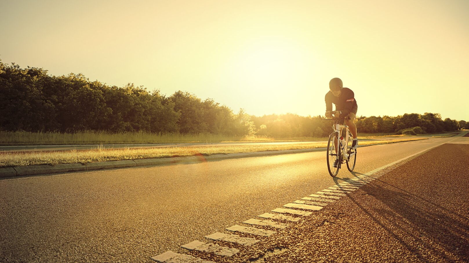 Ein Rennradsportler fährt auf einer Straße, im Hintergrund geht die Sonne unter.