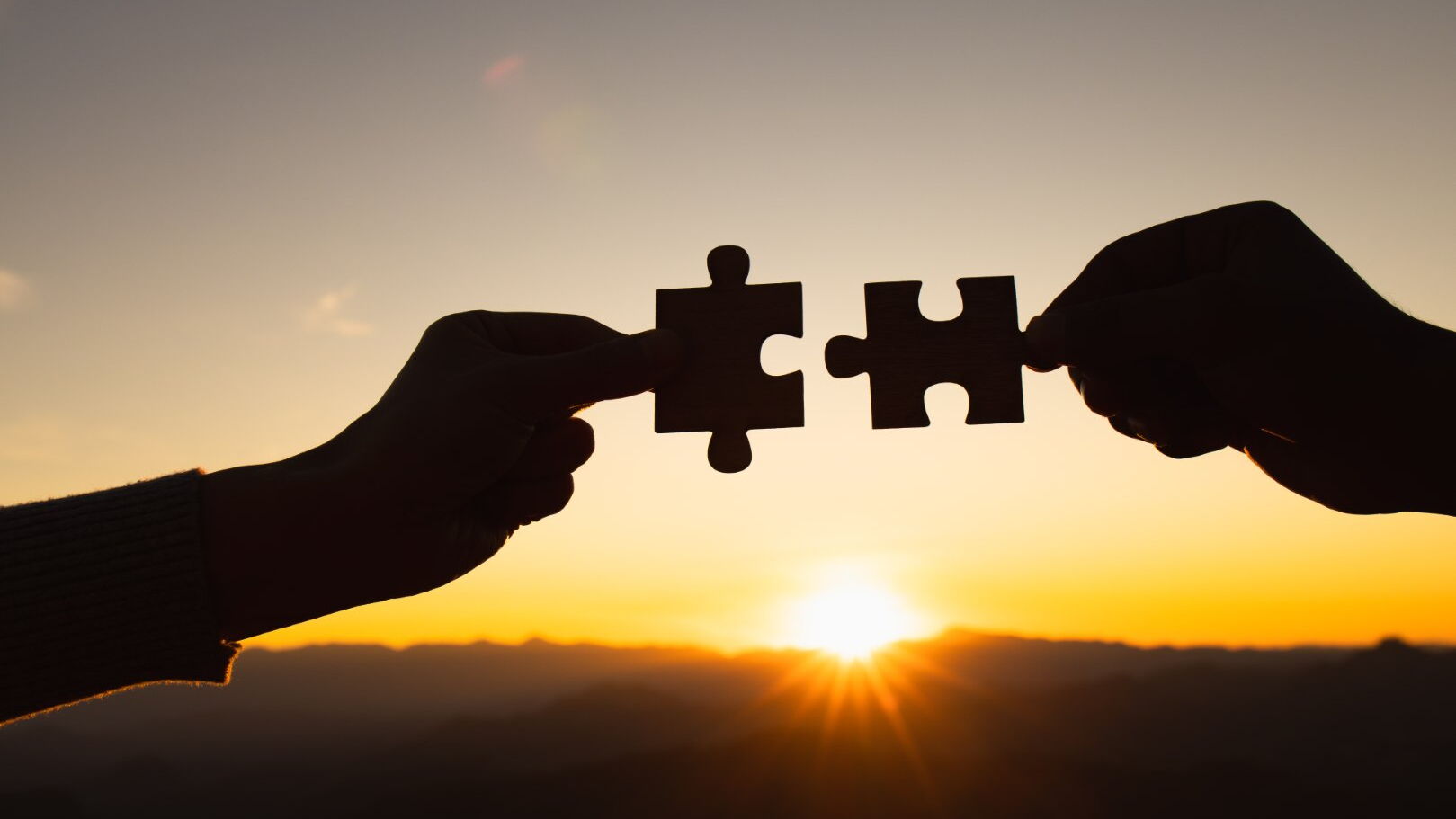 Zwei Hände führen vor einem Sonnenuntergang Puzzleteile zusammen