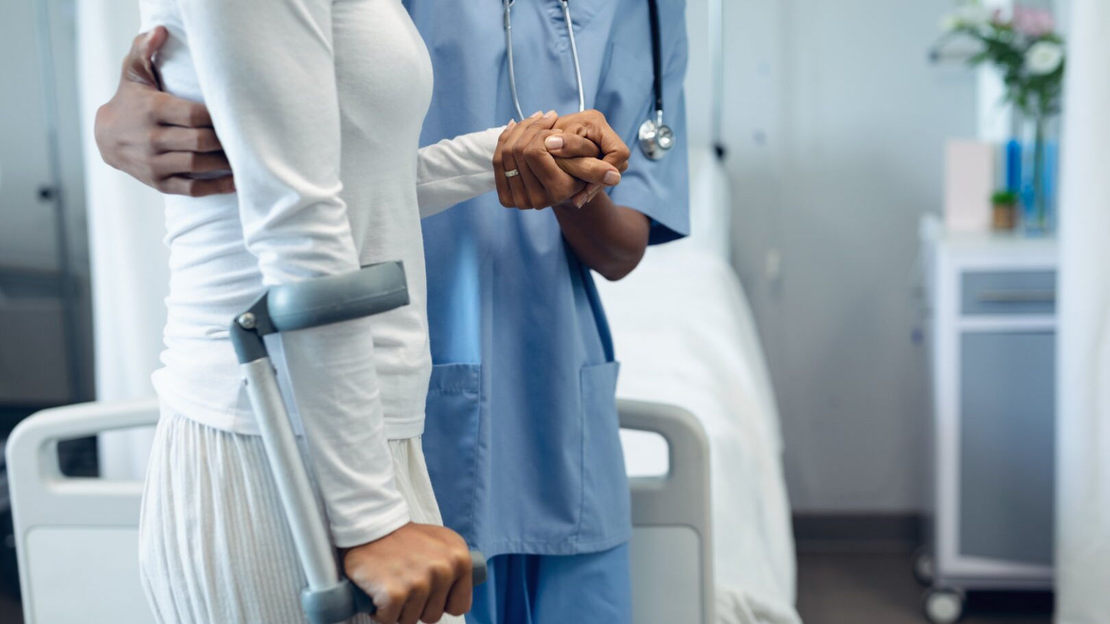 Eine Patientin wird im Krankenhaus von einer Pflegekraft geführt. Sie muss sihc zusätzlich auf einen Gehhilfe stützen.