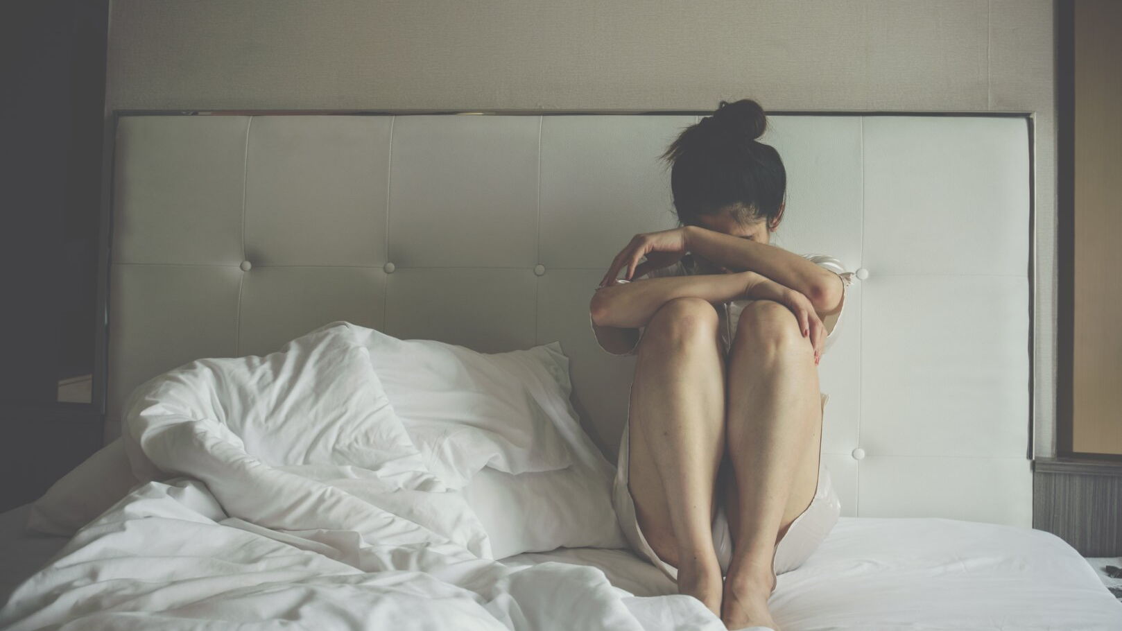 Eine Frau sitzt auf einem Bett und verbirgt ihr Gesicht in den verschränkten Armen.