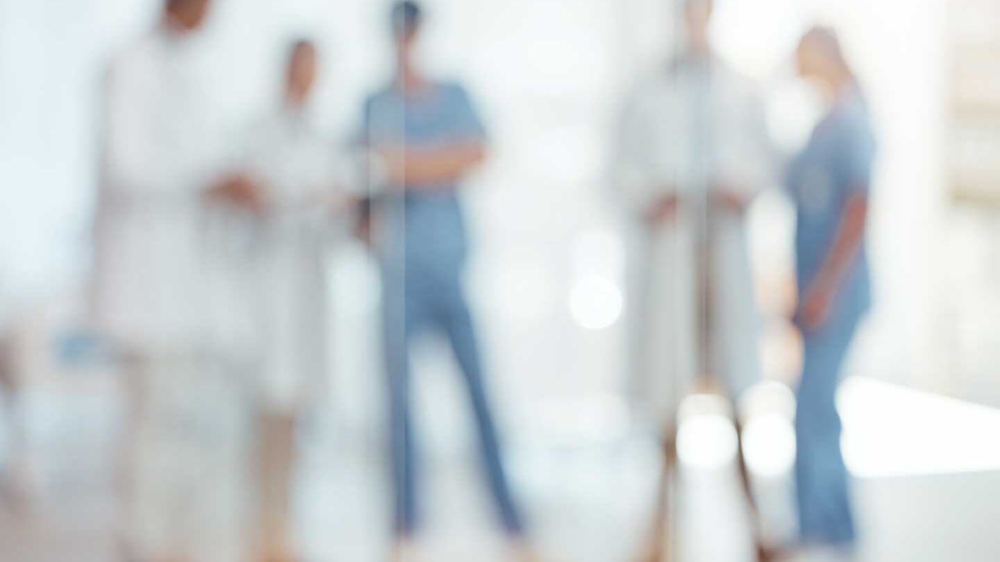 Ein unscharfes Foto von 5 Personen vor einem hellen Hintergrund. Zwei Personen tragen die für Krankenpfleger typische blaue Kluft, drei Personen helle, hüftlange Kittel.