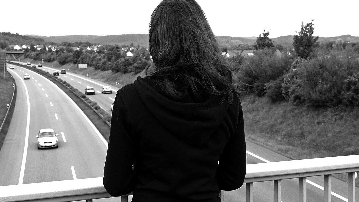 Schwarzweißfoto einer Frau. Die Person wendet dem Betrachter den Rücken zu. Sie steht auf einer Brücke über einer vierspurigen Straße.
