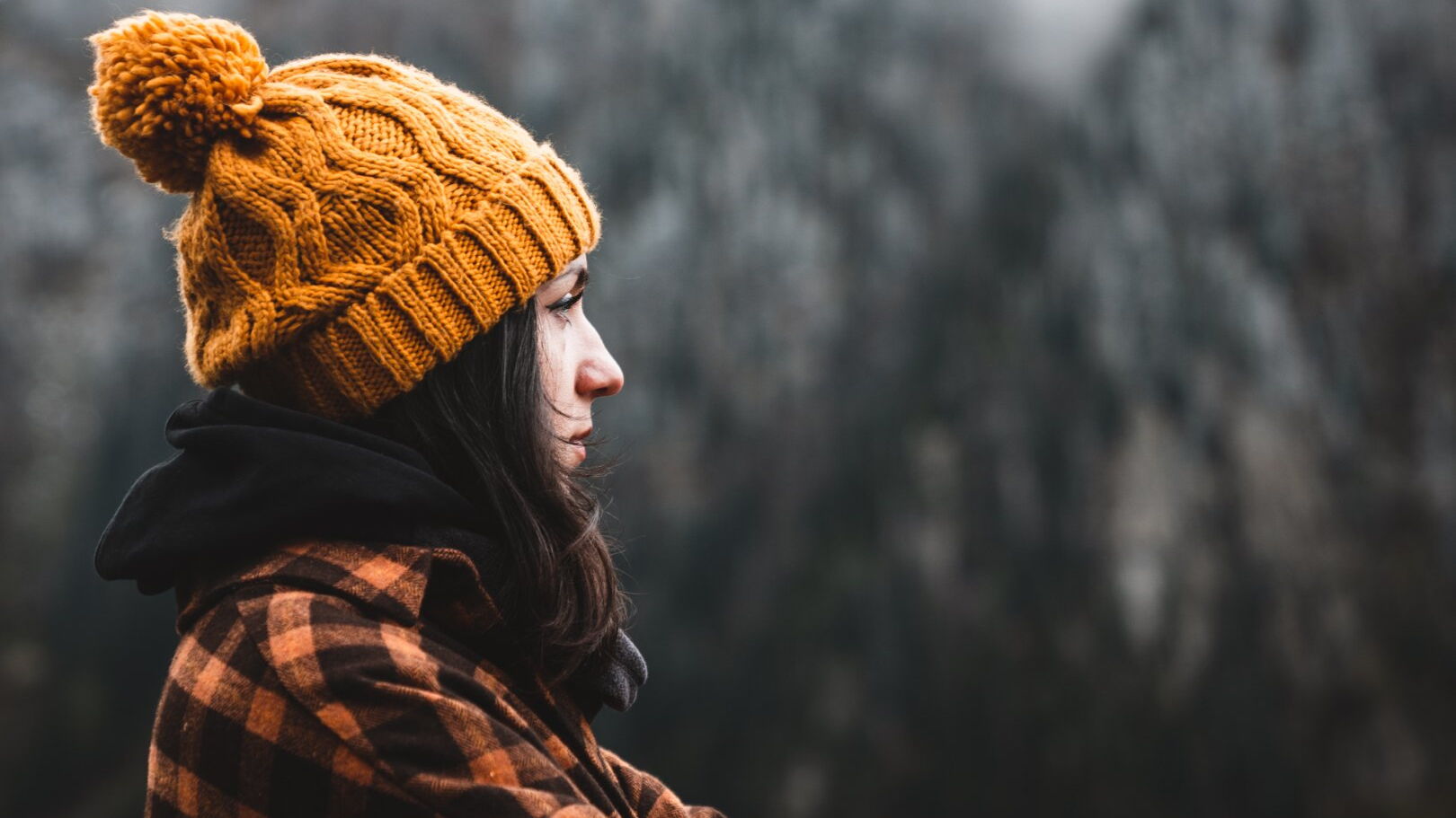 Seitenansicht auf eine nachdenklich in die Ferne schauende junge Frau mit Mütze. Im Hintergrund lässt sich eine Winterlandschaft erahnen.