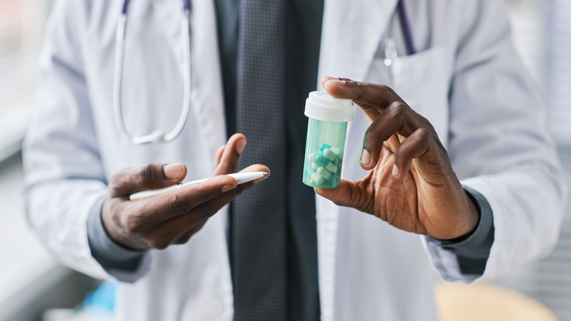 Das Bild zeigt eine Ärztin oder einen Arzt im weißen Kittel mit Stethoskop, die oder der eine grüne Medikamentendose mit weißen und blauen Kapseln in der Hand hält. Mit der anderen Hand wird auf die Dose gezeigt, vermutlich um etwas zu erklären. Der Fokus