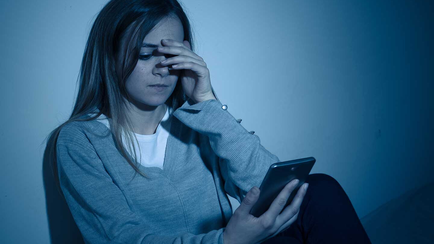 Eine junge Frau sitzt in einem dunklen Raum auf dem Boden. Sie blickt ernst auf ein Smartphone in der rechten Hand. Die linke Hand hält sie halb vor das gesicht.
