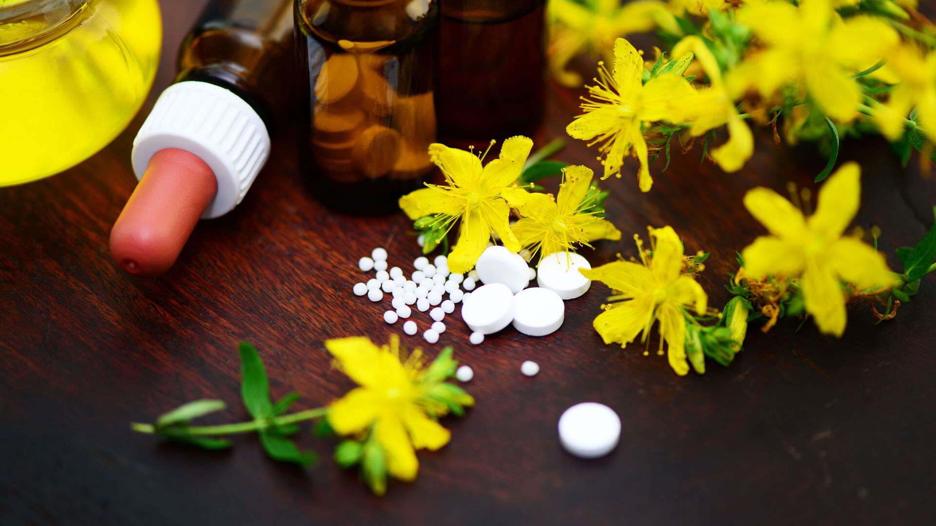 Das Bild zeigt gelbe Johanniskrautblüten neben weißen Tabletten, Globuli und kleinen braunen Fläschchen mit Pipette auf einer dunklen Holzoberfläche. Es symbolisiert natürliche Heilmittel und den Einsatz pflanzlicher Präparate in der Medizin.