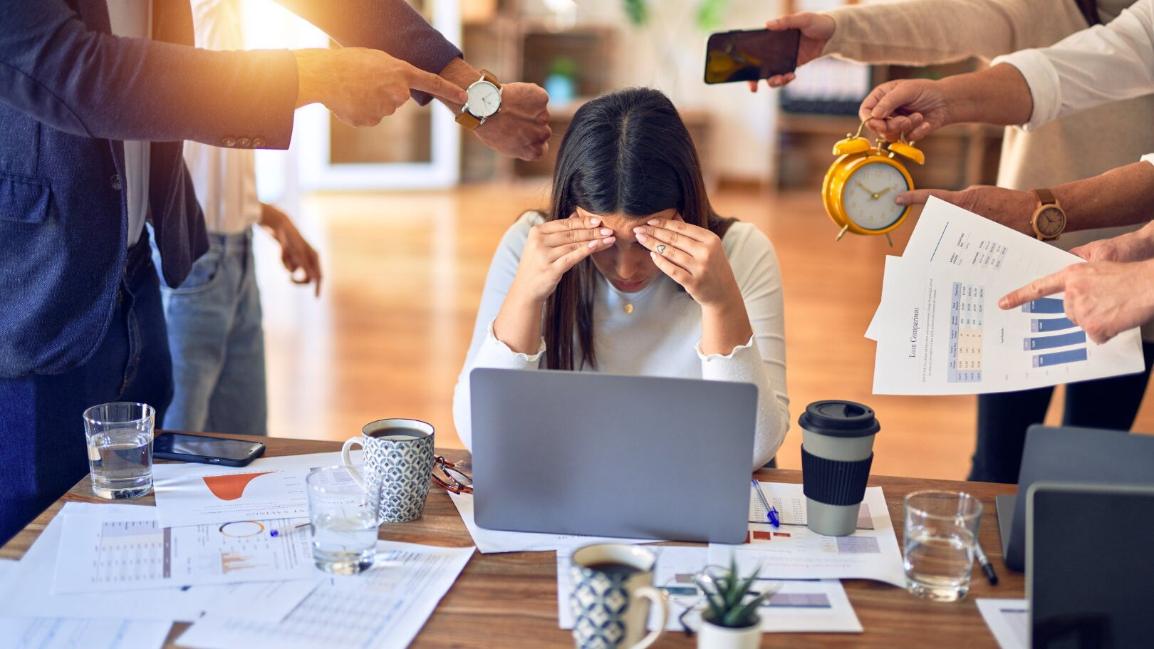 Eine Frau sitzt am Schreibtisch und stützt ihre Stirn auf die Hände. Ihr werden von allen Seiten neue Aufgaben gereicht, was ihr großen Stress bereitet.