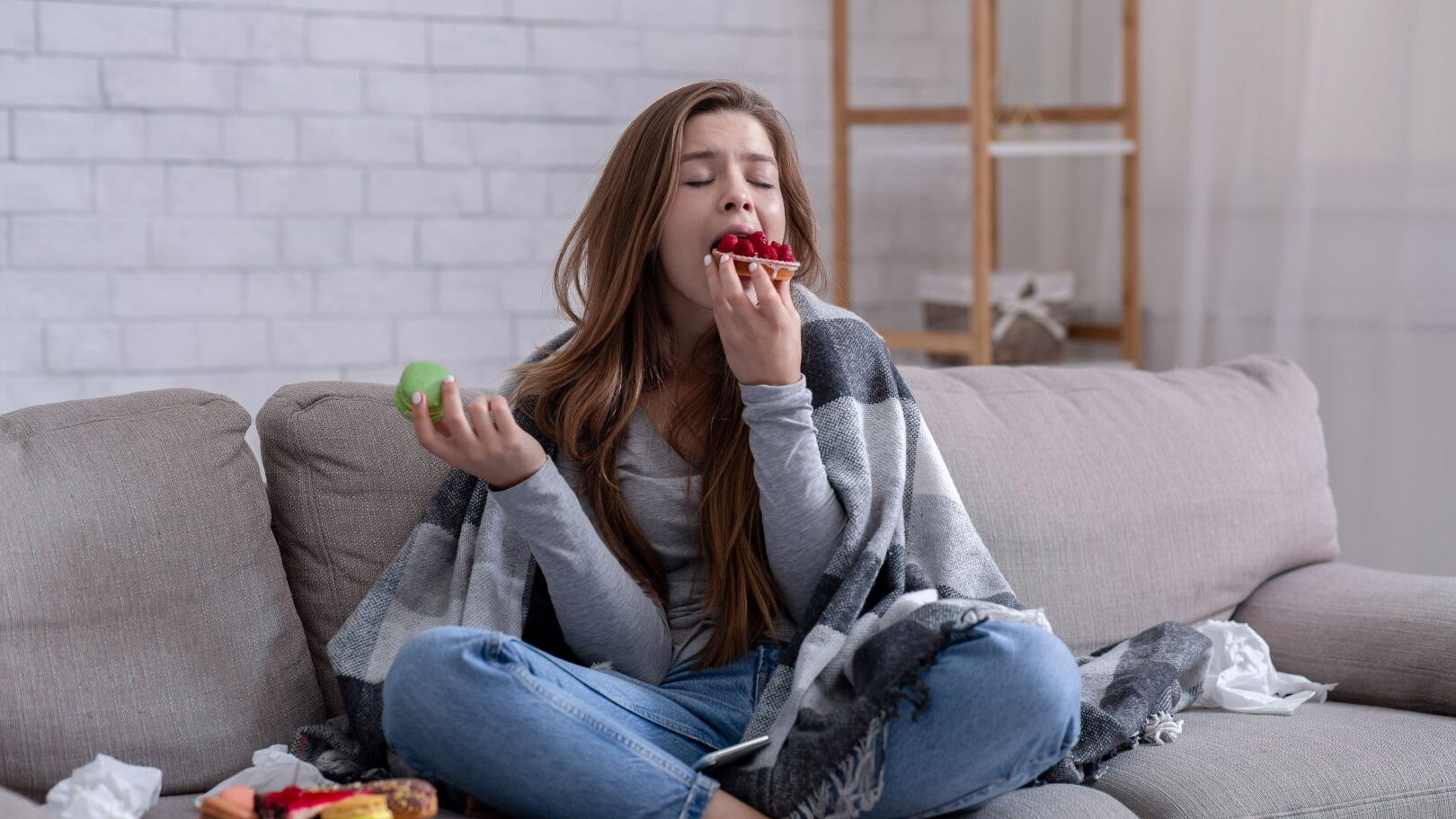 Eine Frau sitzt auf einem Sofa und hat sich eine Decke um die Schultern gelegt. Sie beißt herzhaft in ein Stück Kuchen, weitere Süßigkeiten stehen auf einem Teller auf dem Sofa bereit.