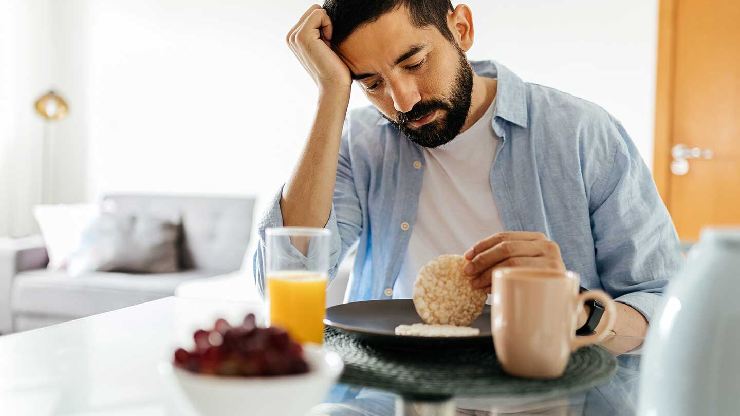 Ein Mann sitzt an einem Tisch, vor sich einen Teller, eine Tasse, ein Glas Saft und eine (unscharfe) Schale mit roen Früchten. Der Mann hat den Kopf auf die rechte Hand gestützt und schaut auf eine Reiswaffel in seiner linken Hand.
