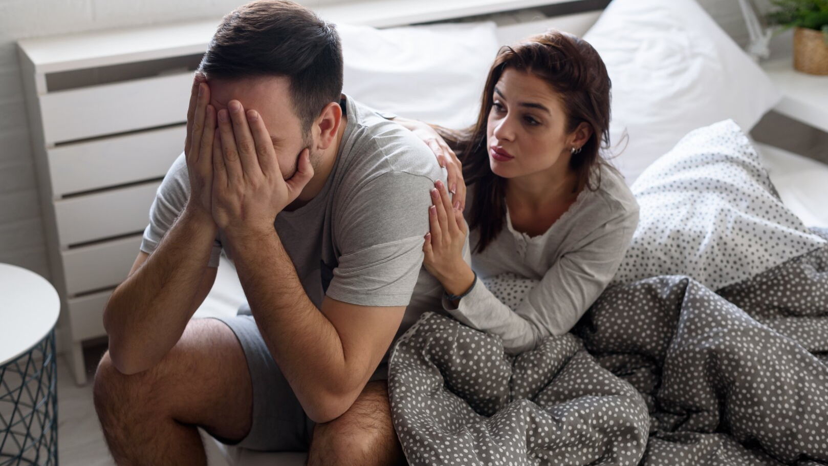 Ein Mann hält sich das Gesicht mit beiden Händen, während er auf der Bettkante sitzt. Seine Partnerin versucht ihm dahinter Trost zuzusprechen.