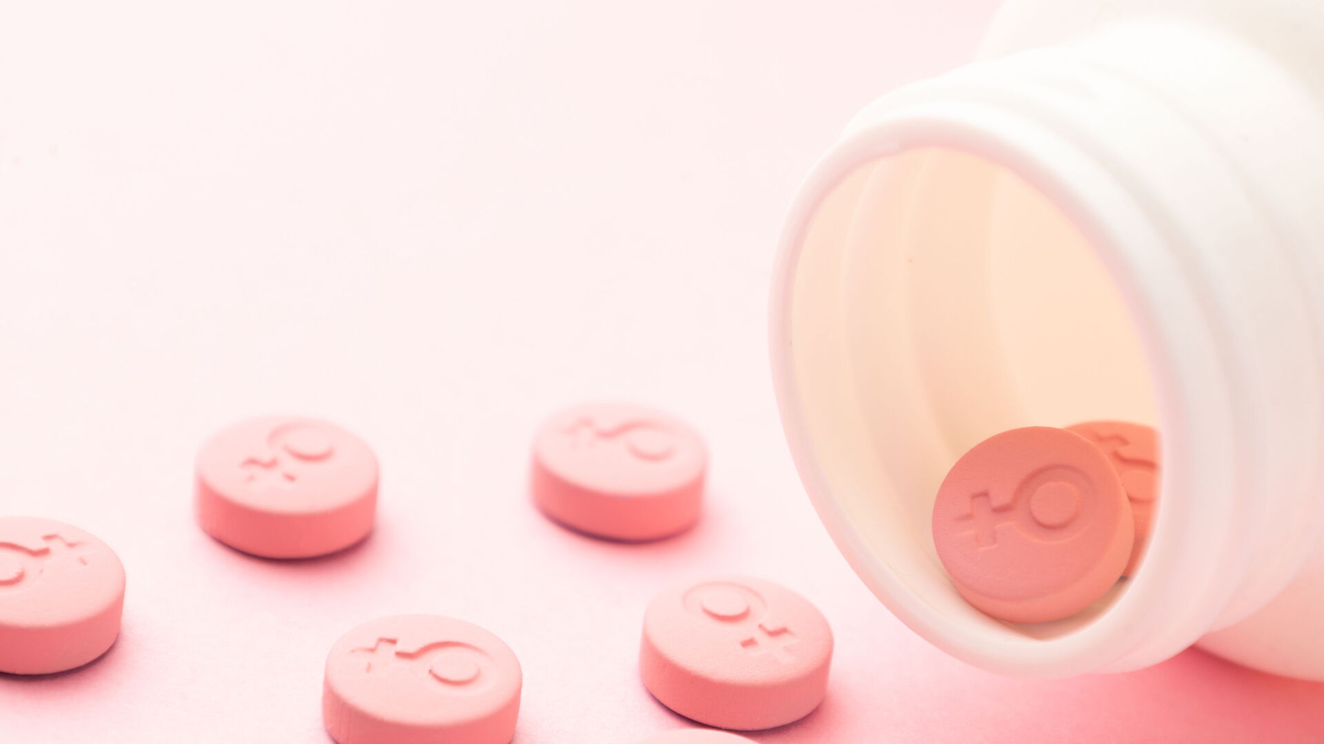 Das Bild zeigt eine offene weiße Medikamentenflasche, aus der mehrere rosafarbene Tabletten auf eine rosa Oberfläche gerollt sind. Auf den Tabletten ist ein Symbol eingraviert, das das weibliche Geschlechtssymbol darstellt.